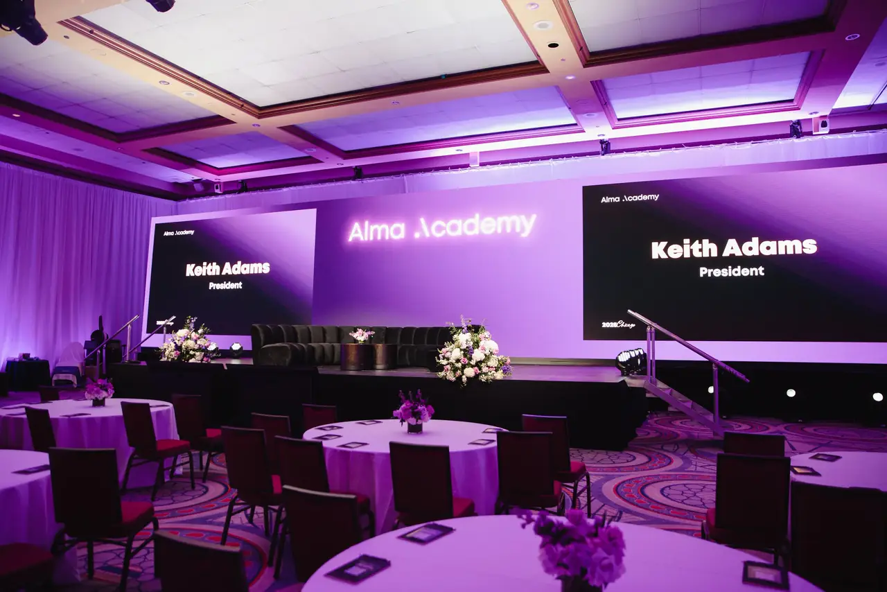 Alma Academy ballroom set for a general session: round tables, purple uplighting and three LED screens